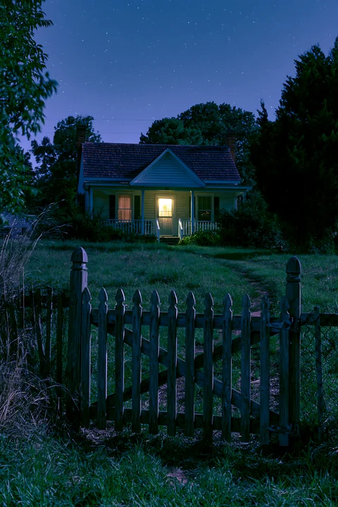 A country home stands illuminated with a warm light at night, surrounded by tall grass and trees.