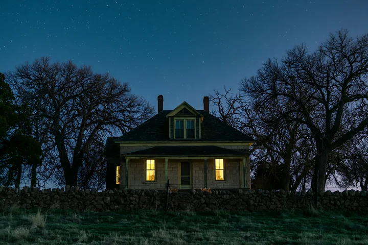 A country house stands under a starry sky, bright lights glowing from the windows.