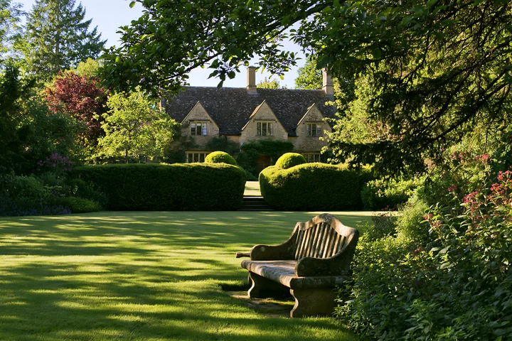 A country house surrounded by hedges sits in a garden with a bench.