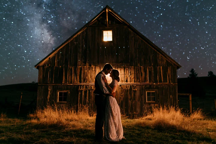 A couple dancing under stars outdoors stands in front of a rustic barn.