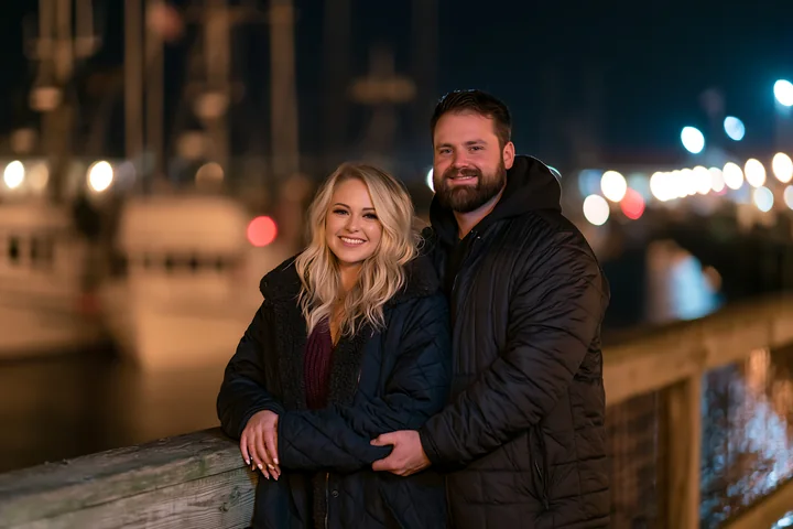 A couple holding hands beneath the night sky stands on a wooden dock at a marina.