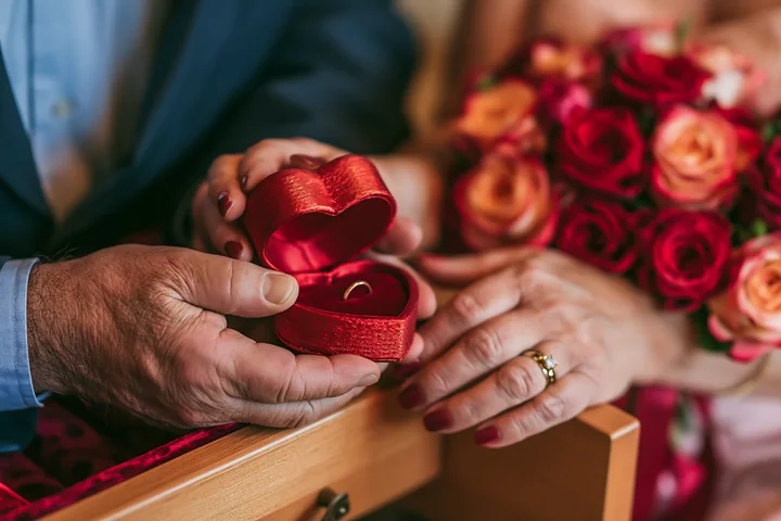 A couple is holding a heart-shaped ring box with a ring inside while surrounded by roses.