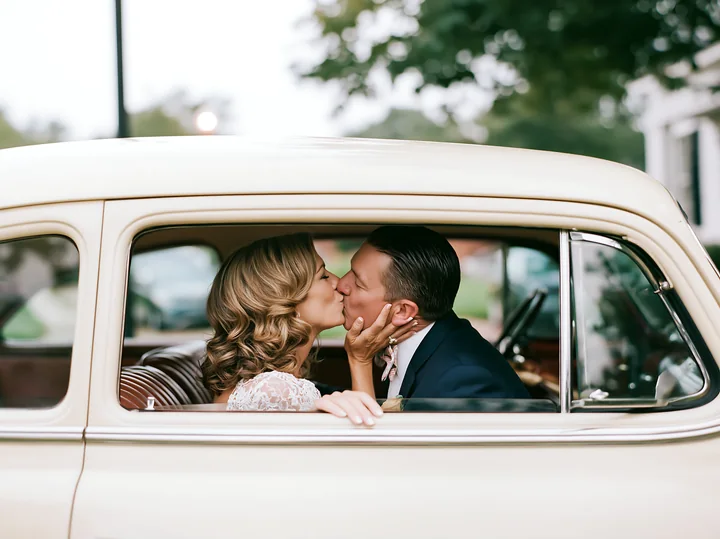 A couple is kissing inside a vintage car, showing affection and joy.