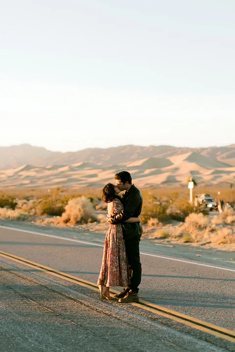 A couple is kissing on a quiet road surrounded by desert terrain at sunset.