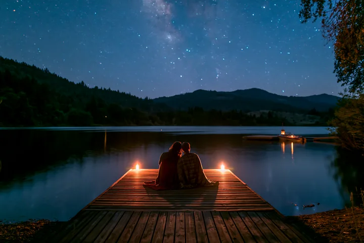A couple is watching stars beside a lake, sitting on a wooden dock with candles lit around them.