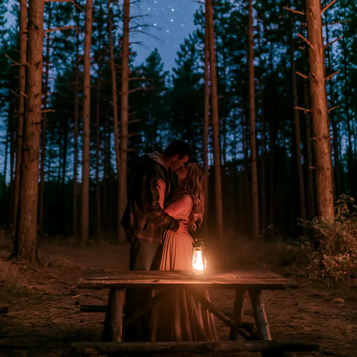 A couple kisses beneath stars beside a glowing lantern in a forest.