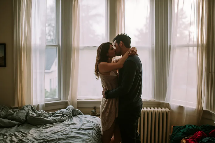 A couple kisses in a bright bedroom filled with natural light.