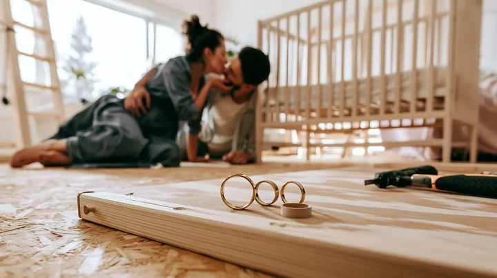 A couple kisses in a cozy room while wedding rings rest on a wooden surface.