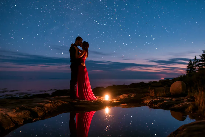 A couple kisses on a rocky shore under a starry night sky, with a gentle glow from a candle reflecting in the water.