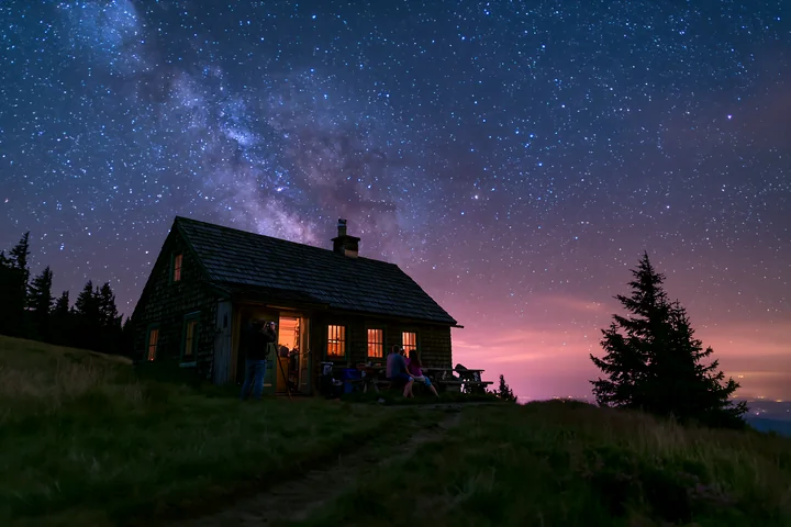 A couple photographing Milky Way at night near a cabin surrounded by trees.
