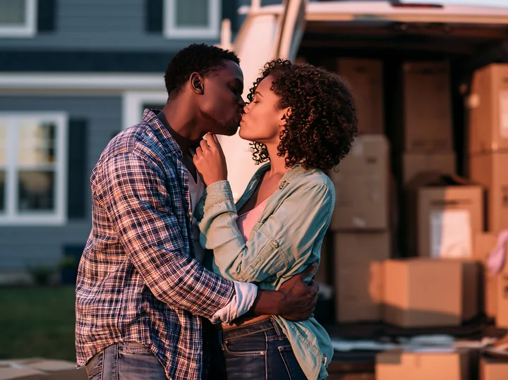 A couple shares a kiss while surrounded by moving boxes outside their new home.