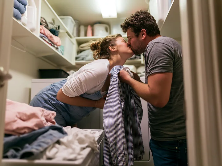 A couple shares a tender kiss while standing in a laundry room filled with clothes.