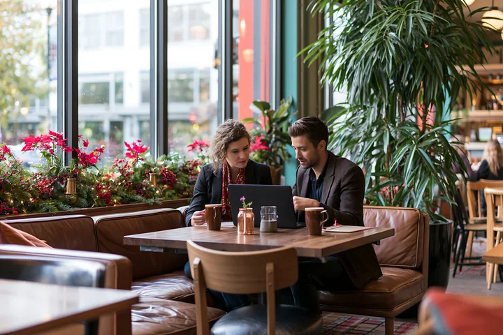 A couple sits at a wooden table in a coffee shop, working on a laptop.