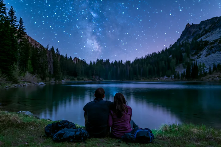 A couple sits by a lake, admiring the starry night sky filled with bright stars.