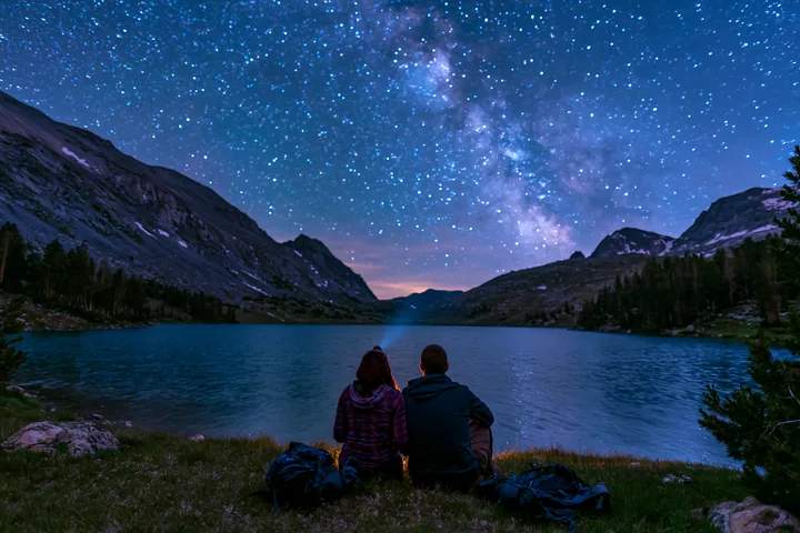 A couple sits by a lake while looking up at a starry night sky filled with stars.