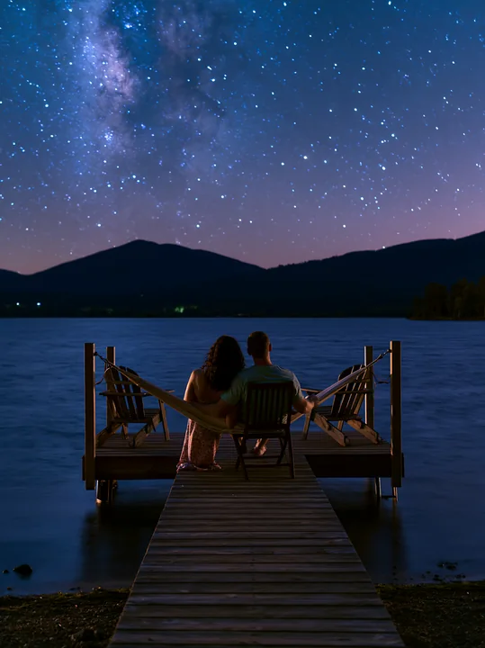 A couple sits in a hammock on a dock by the lake, surrounded by mountains and a clear night sky filled with stars.