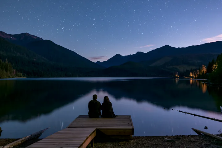 A couple sits on a dock under stars near a mountain lake, enjoying the night sky reflected in the calm water.