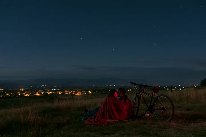A couple sits on the grass at night, wrapped in a blanket, sharing a kiss under the stars.