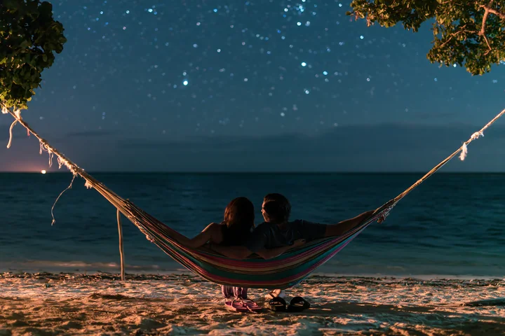 A couple sitting in a hammock under stars relaxes together on the beach, with the ocean in the background.