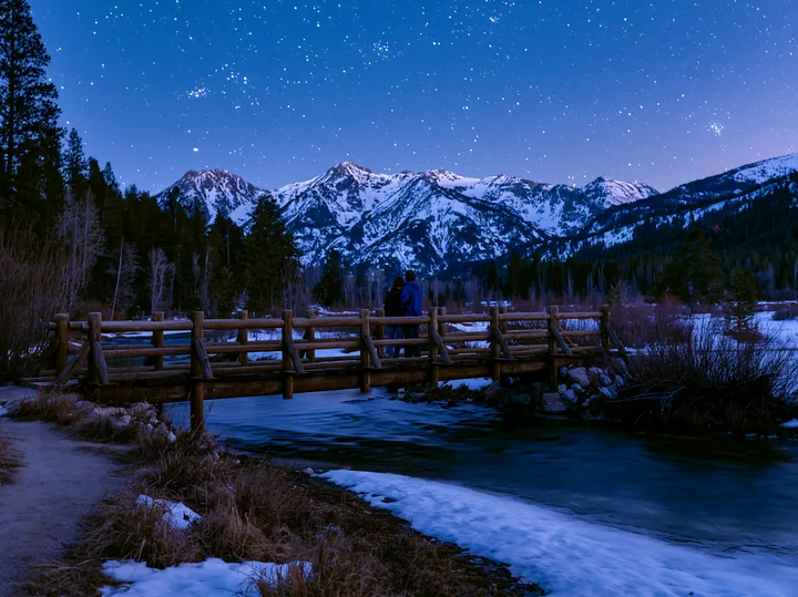 A couple stands on a wooden bridge, gazing at a star-filled sky above snowy mountains.