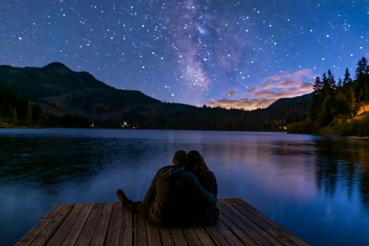 A couple under stars near a mountain lake relaxes on a wooden dock.