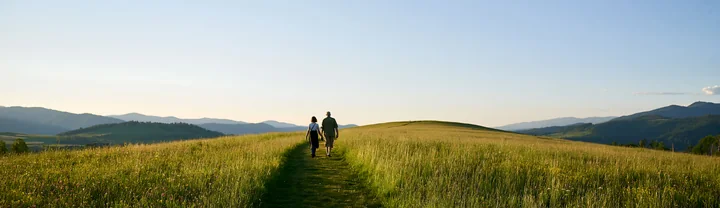 A couple walks along a grassy path in a meadow, surrounded by hills and mountains, enjoying the clear sky.