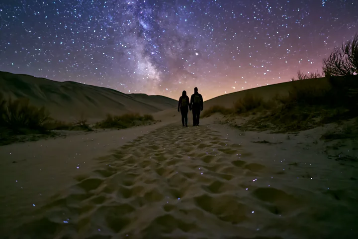 A couple walks on a sandy path in a desert under a bright starry sky.