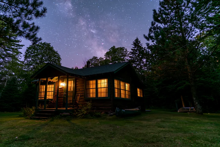 A cozy holiday home under stars stands in a forest, with warm lights glowing from its windows.