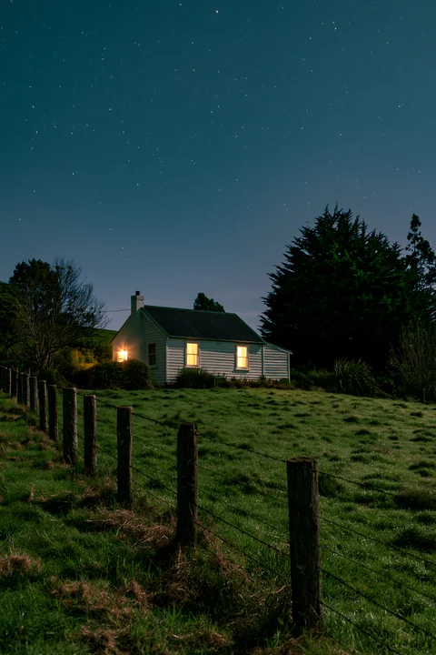 A cozy house in a meadow beneath stars stands surrounded by grass and trees.