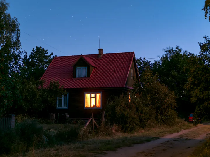 A cozy house stands illuminated at dusk, surrounded by trees and shrubs.