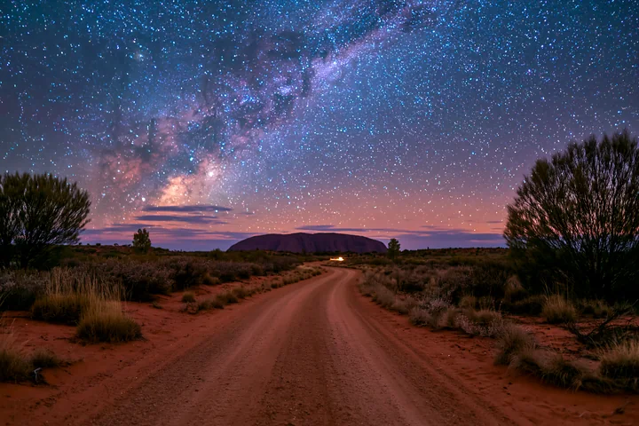 A dirt road leads to Uluru under a starry night sky filled with bright stars and the Milky Way.