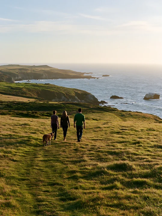 A family with a dog walks along a path on a coastal meadow under a broad open sky.