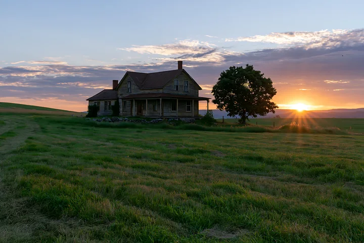 A farmhouse sits in a green field as the sun sets in the background, creating a warm glow in the sky.