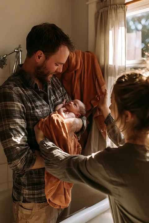 A father holds a wrapped newborn while a mother prepares a towel in a bathroom.