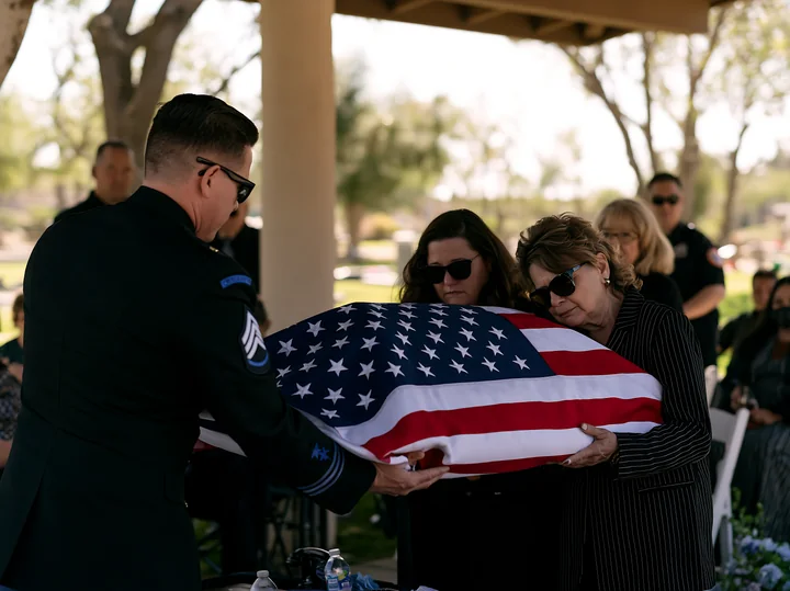 A folded American flag presented to a grieving family at a funeral service, with mourners seated in the background under a shaded area.