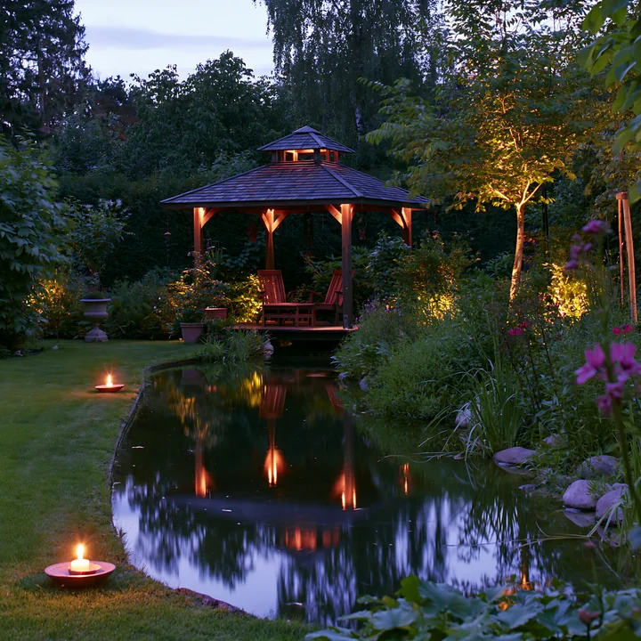 A garden pavilion is lit softly in the evening, surrounded by greenery and a small pond reflecting warm lights.