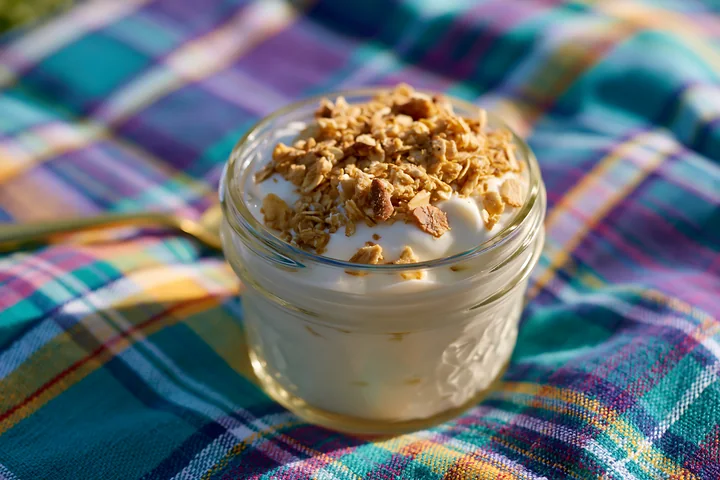 A glass jar filled with cream dose with oat flakes sits on a colorful plaid cloth in natural light, emphasizing the textures of both the cream and the oat toppi