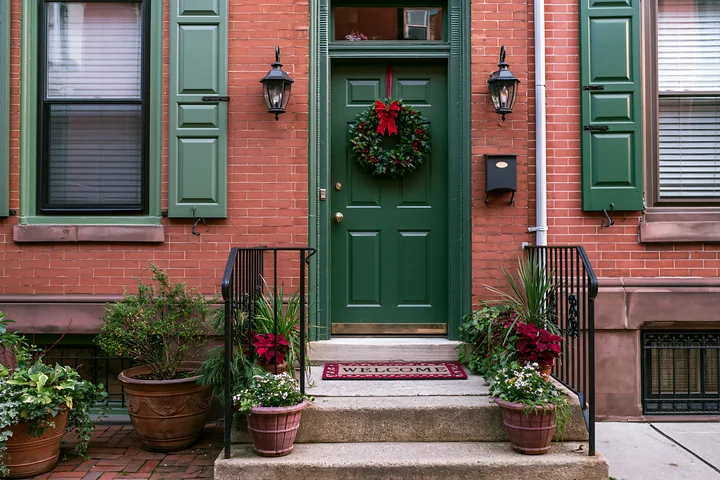 A green front door features a wreath and a welcome mat at the entrance of a brick home, surrounded by potted plants and a well-maintained walkway.