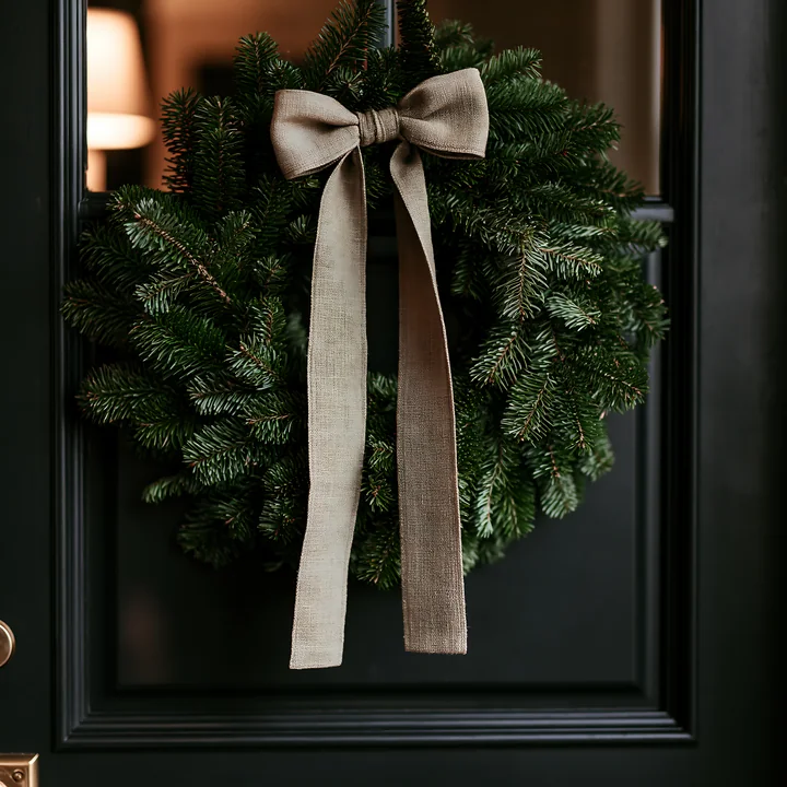 A green wreath adorned with a beige bow hangs on a dark front door.