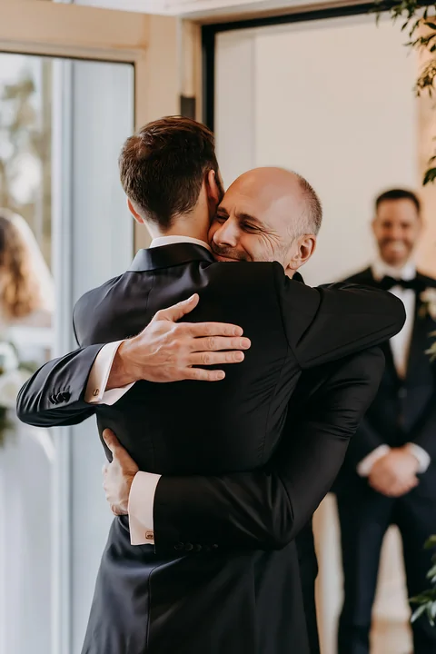 A groom and his best man share an emotional wedding ceremony embrace in a bright indoor setting.