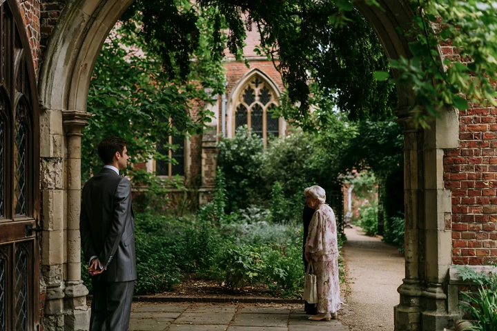 A groom stands near a church arch, waiting while an elderly woman approaches.