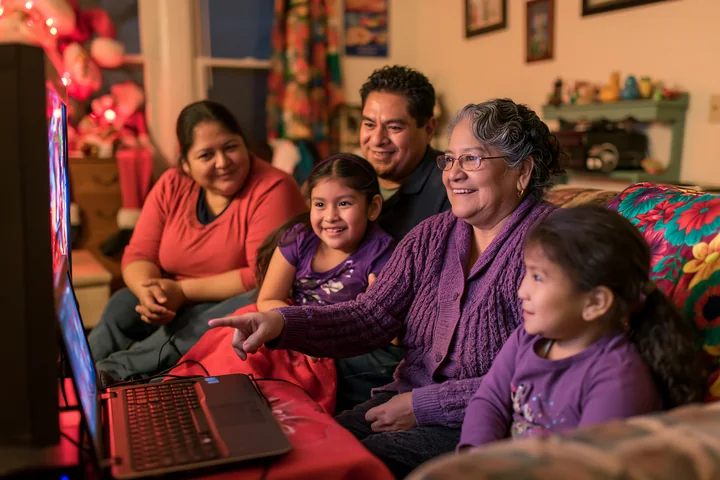 A group gathers in a warm living room, focused on a screen while watching a movie.