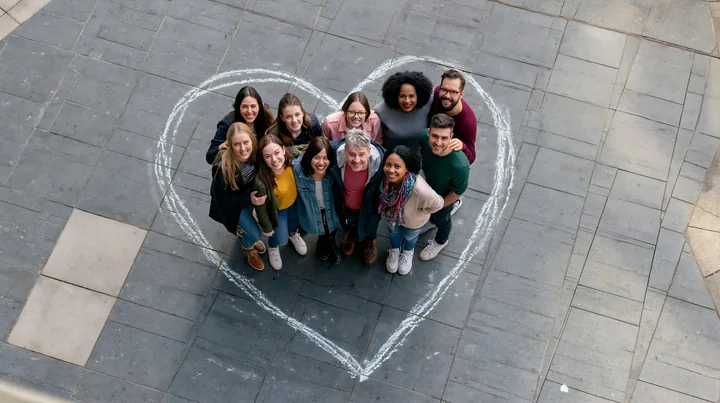 A group of diverse people stands together inside a heart-shaped chalk outline on pavement in an outdoor setting.