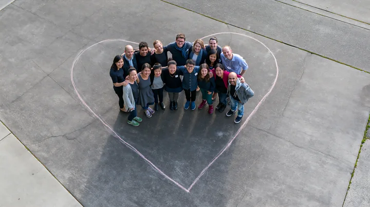 A group of people stands inside a large heart-shaped chalk outline on pavement, smiling for a cheerful outdoor campaign.