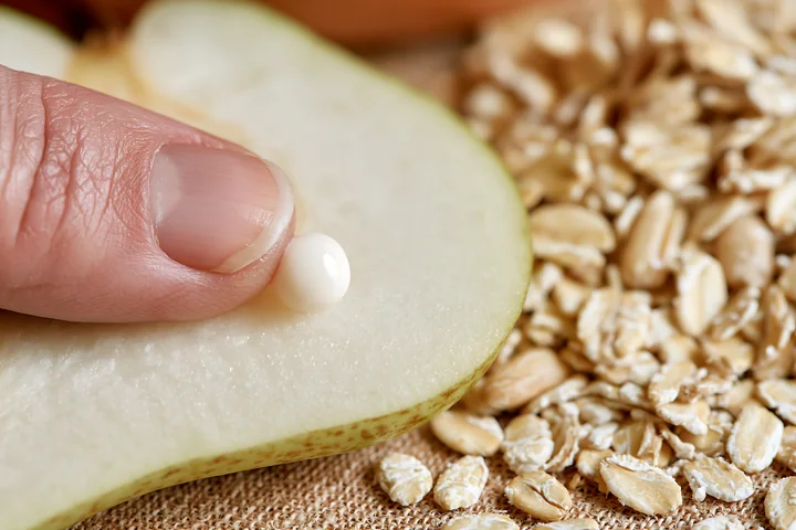 A hand applies a small cream dose with natural skincare ingredients to a slice of pear next to oats on a textured surface.