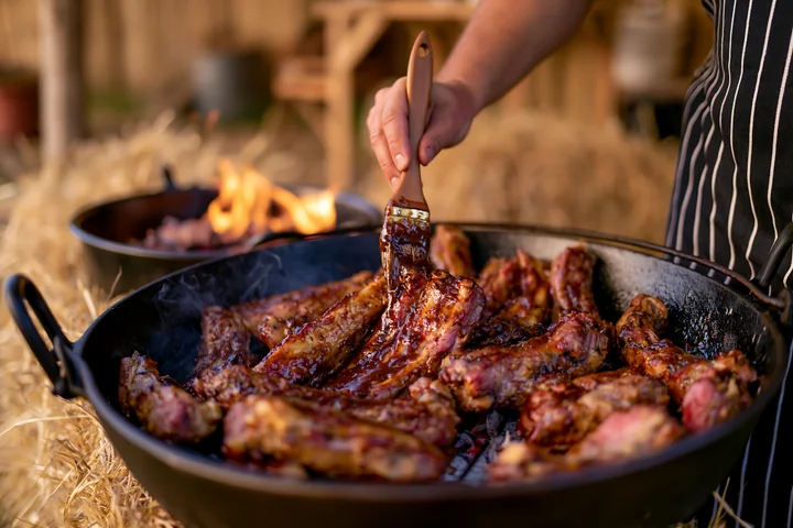 A hand brushes marinade onto goat meat being cooked over an open flame at a countryside festival.
