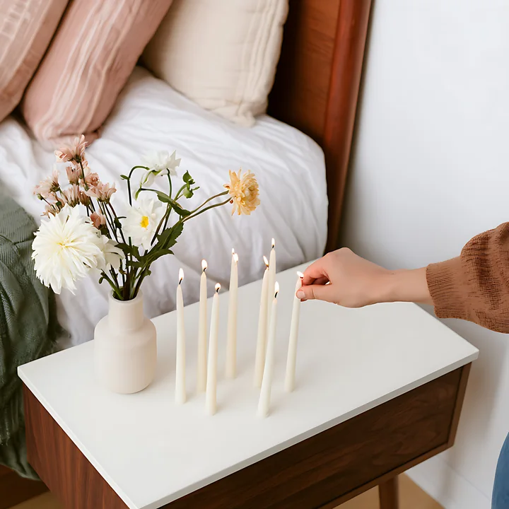 A hand lights candles on a bedside table with a vase of flowers.