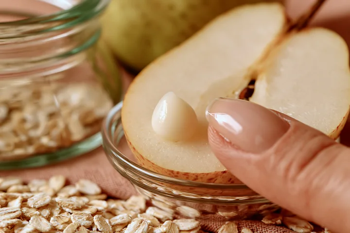 A hand places a cream dose with natural skincare ingredients on a slice of pear surrounded by oats and a green fruit.