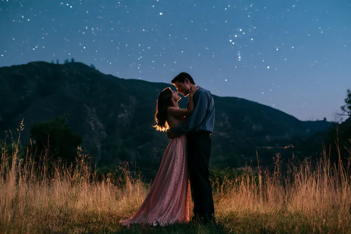 A happy couple kissing beneath stars in nature during the night with mountains in the background.