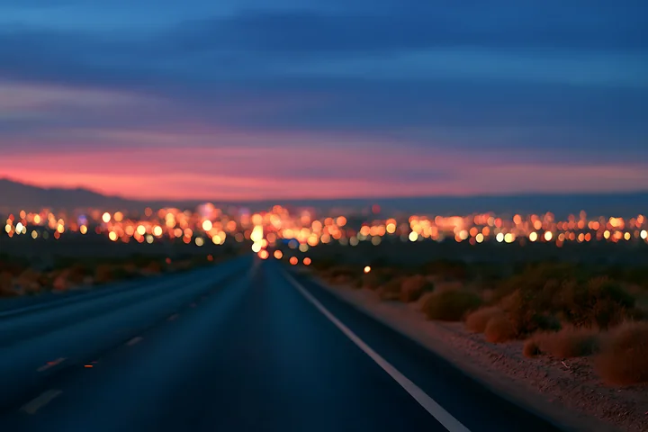 A highway stretches towards a city in the distance as dusk falls, with bright lights illuminating the urban landscape.